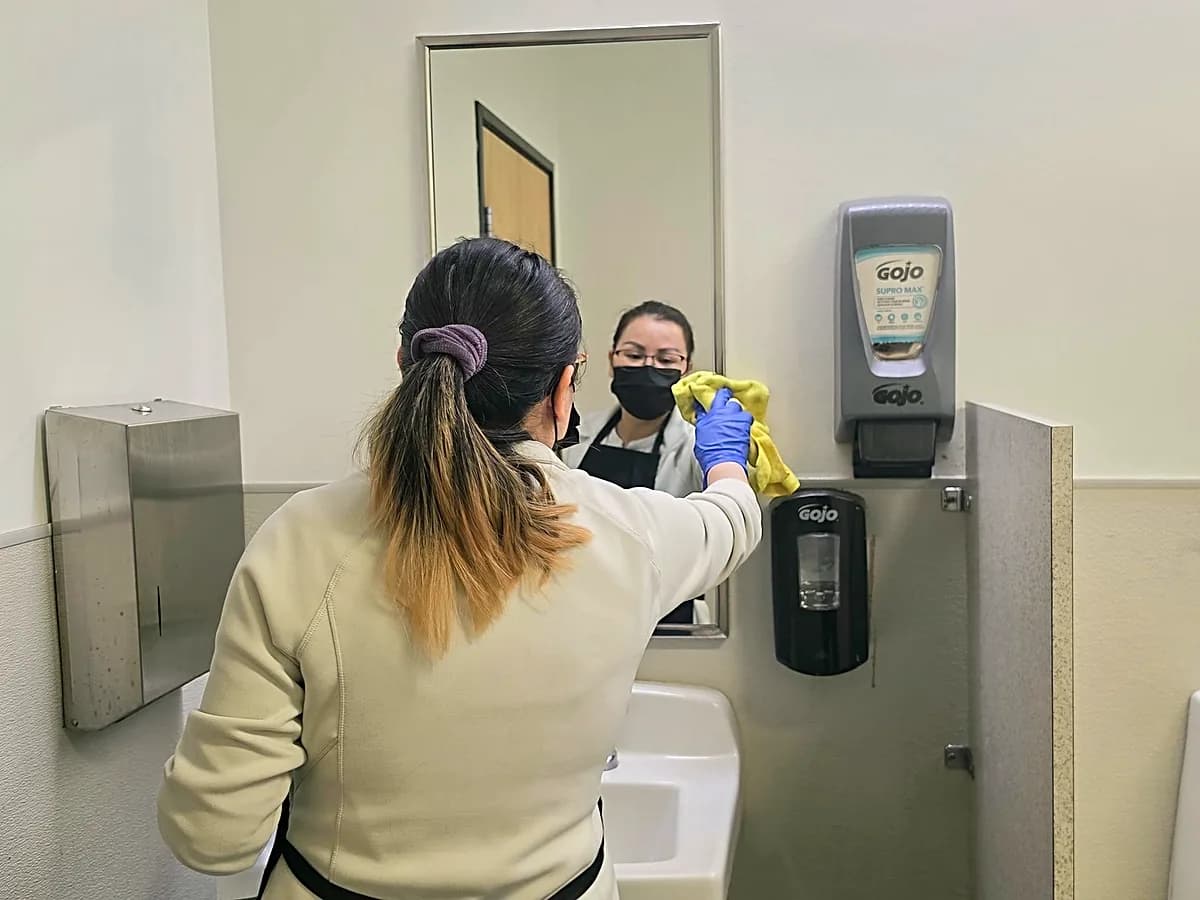 Janicat cleaner wiping a restroom mirror above the sink
